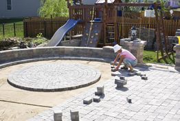 Person in a white hat laying pavers in a backyard with a playset in the background.
