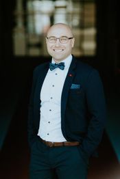 Ein lächelnder Mann mit Brille und blauer Fliege in Anzug posiert in einem dunklen Raum.