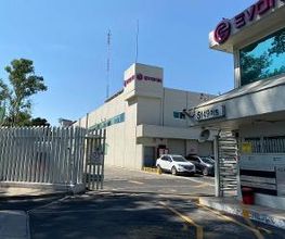 Edificio blanco con logos de empresa y estacionamiento, visto desde el portón de entrada.