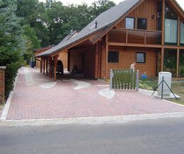Holzhaus mit Doppelgarage und gepflasterter Auffahrt, umgeben von Bäumen und ruhiger Straße.