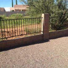 Gravel driveway beside a metal fence with trees and houses in the background on a sunny day.