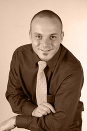 Ein lächelnder Mann in Hemd und Krawatte, mit gekreuzten Armen, vor hellem Hintergrund.