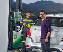 Persona sonriente en gasolinera, otra persona surtiendo combustible. Montañas al fondo.