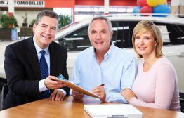 A man with a clipboard helps a couple signing papers at a car dealership table.