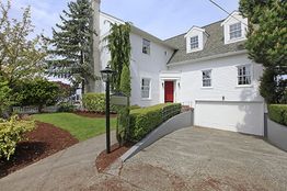 White two-story house with red door, driveway, and green lawn surrounded by trees and bushes.