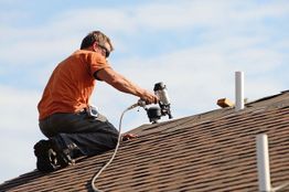 A man in an orange shirt uses a nail gun while working on a shingled roof.