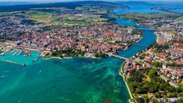 Stadt am Bodensee mit Brücke und Hafen, umgeben von grünem Land und blauem Wasser. Luftaufnahme.