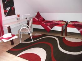 Zimmer mit zwei Betten, Schreibtisch, Stuhl und Teppich in Rot- und Brauntönen.
