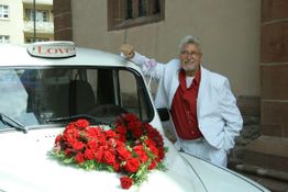 Ein Mann in weißem Anzug lehnt an einem mit Rosen geschmückten Auto mit "Love"-Schild.