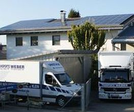Transporter und LKWs stehen vor einem Gebäude mit Solarpanels, umgeben von Bäumen.