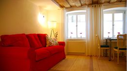 Gemütliches Zimmer mit rotem Sofa, Holzboden, Schreibtisch und zwei Fenstern mit Vorhängen.