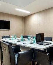 Sala de reunião com mesa retangular, cadeiras pretas e TV na parede.