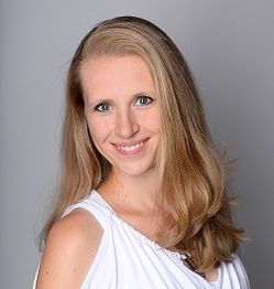Eine lächelnde Frau mit blonden Haaren trägt ein weißes Oberteil vor grauem Hintergrund.