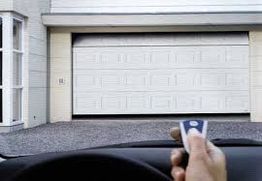 Hand holding a remote, opening a white garage door in a driveway.
