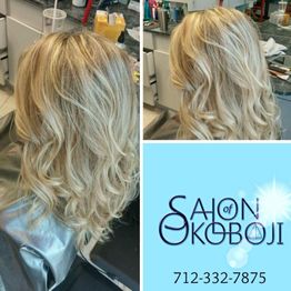 Blonde hair with loose curls at a salon; logo and contact number displayed on the bottom right.