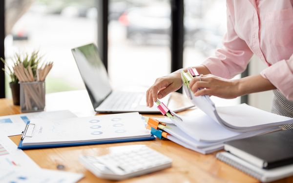 Eine Frau blättert durch viele zusammen geheftete Dokumente, die auf ihrem Schreibtisch liegen.