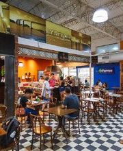 Cafeteria movimentada com pessoas em mesas e um balcão de atendimento ao fundo. Chão xadrez.