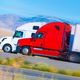 Red and white trucks driving side by side on a highway in a mountainous landscape.