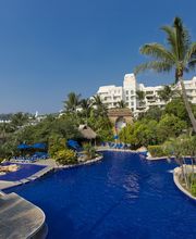 Piscina azul con palmeras, sillas azules y un hotel blanco de fondo bajo un cielo despejado.