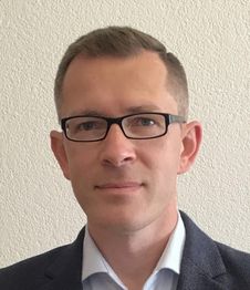 Ein Mann mit Brille und blauem Anzug vor einer weißen Wand.
