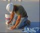 Worker kneeling on a surface, using a brush with a white bucket and blue cloth nearby.