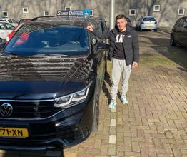 Een man staat naast een zwarte auto met een kenteken, op een zonnige dag.