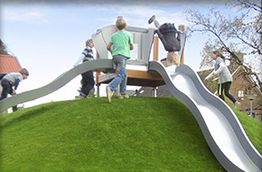 Kinder spielen auf einem Hügel mit zwei Rutschen bei sonnigem Wetter.