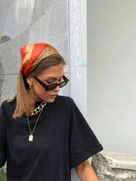 Woman in sunglasses and scarf, wearing a black shirt and necklaces, stands by a gray wall.