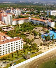 Vista aérea de un complejo hotelero junto a la playa, con piscinas y palmeras.