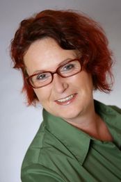 Eine lächelnde Frau mit rotem Haar und Brille in grüner Bluse vor grauem Hintergrund.