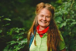 Eine Frau steht lächelnd im Wald, trägt ein grünes Hemd und ein rotes Halstuch.