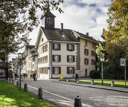Straßenszene mit historischem Gebäude und Uhrenturm bei Herbstwetter. Personen spazieren entlang.