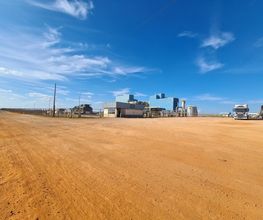 Estrada de terra com edifício industrial ao fundo e caminhões estacionados, sob céu azul.