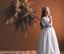 Niña vestida de blanco con corona de flores, junto a un ramo seco en una habitación de madera.