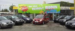 Autohändler mit vielen Autos im Freien, zentral ein rotes Auto. Großes Banner mit buntem Design.
