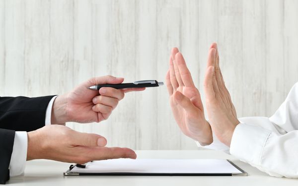 Ein Mann hält einer Kundin symbolisch den Kugelschreiber, zum Unterschreiben des vorliegenden Vertrags hin. Diese lehnt durch zwei erhobene Hände abwehrend ab.