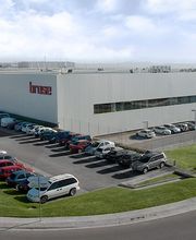 Edificio industrial moderno con estacionamiento lleno y césped verde bajo cielo azul.