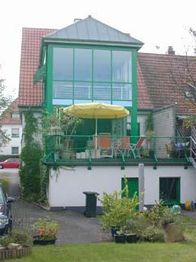 Zweistöckiges Haus mit Balkon, gelbem Sonnenschirm und Flachdach-Wintergarten, grüne Fensterrahmen.