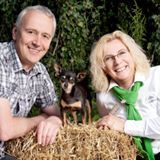 Ein Mann, eine Frau und ein kleiner Hund posieren lächelnd hinter einem Strohballen im Freien.