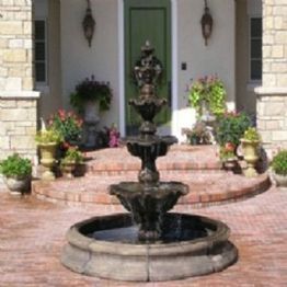 A stone fountain in front of a house with brick steps and potted plants near the entrance.