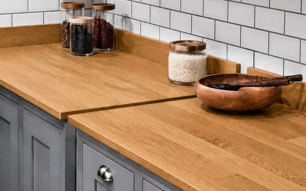 Encimera de cocina de madera maciza o chapada en tono natural, con vetas visibles y acabado cálido, acompañada de tarros de vidrio con alimentos y un bol de madera sobre muebles blancos y salpicadero de azulejos.