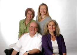 Vier lächelnde Personen posieren zusammen vor einem neutralen Hintergrund.