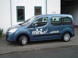 Blauer Minivan mit der Aufschrift "minicar" auf der Seite, geparkt vor einem Gebäude.