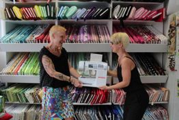 Zwei Personen freuen sich in einem Stoffladen über eine Nähmaschine. Bunte Stoffe im Hintergrund.
