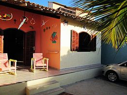 Casa com varanda, duas cadeiras, decoração colorida e carro estacionado ao lado, sob sol tropical.