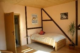 Ein einfaches Zimmer mit Holzbett, Dekokissen, Topfpflanze und schrägen Holzbalken an der Wand.