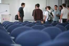 Leere blaue Stuhlreihen im Vordergrund, Gruppe von Menschen stehend im Hintergrund.