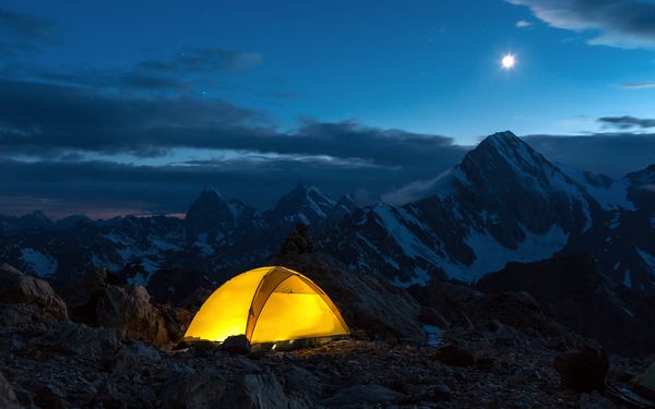 Ein hell erleuchtetes gelbes Zelt bei Nacht wurde in den Bergen aufgebaut. Im Hintergrund ist ein verschneites Bergpanorama zu sehen.