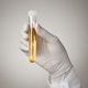 Gloved hand holding a test tube with yellow liquid against a neutral background.