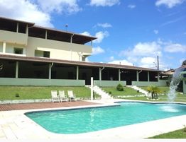 Casa com piscina, cadeiras de sol e céu azul. Jardim bem cuidado e árvores ao fundo.